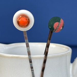 Può includere: Due forcine decorative. Una forcina presenta un bottone bianco con un centro arancione e finiture dorate. L'altra forcina ha un bottone in tessuto verde con un motivo floreale rosa. Entrambe le forcine sono in un contenitore di ceramica bianca su sfondo blu.