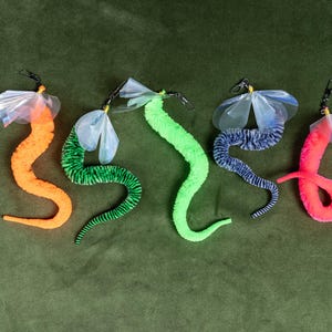May include: Five fuzzy, worm-shaped toys in orange, green, lime, blue, and pink. Each toy features a flexible, textured body and a translucent, iridescent plastic wing attachment at the top, secured with a small black metal swivel clip.