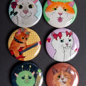 Puede incluir: Un conjunto de seis botones redondos con temática de gatos. Cada botón presenta una ilustración de gato diferente, incluyendo un gato con una rosa, un gato sacando la lengua y un gato tocando la guitarra. Los botones tienen un acabado brillante.