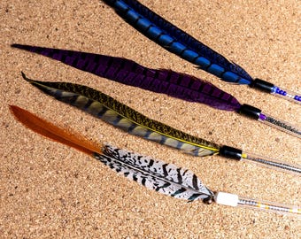 May include: Four decorative feathers attached to clear plastic wands. The feathers are blue, purple, yellow-green with black stripes, and orange with black and white spotted patterns. Each wand contains small colorful beads inside the clear tube.