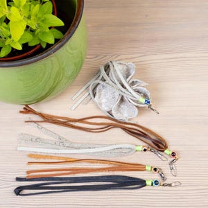 Op de afbeelding: Een groene keramische pot met een plant staat naast verschillende leren kwasten in bruin, grijs en zwart. Elke kwast heeft een kleine kraal en een zilverkleurige sluiting.