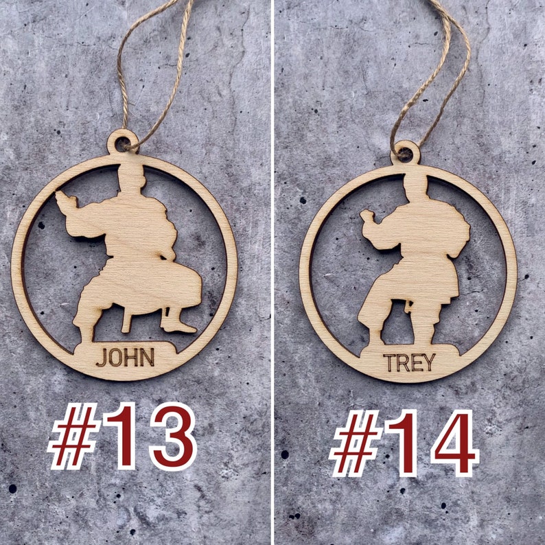 May include: Two wooden ornaments featuring a silhouette of a person in a karate pose. The ornament on the left reads "JOHN #13" and the ornament on the right reads "TREY #14".