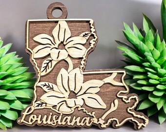 May include: Wooden ornament shaped like the state of Louisiana with a brown wood background and a light wood design of magnolia flowers. The word "Louisiana" is written at the bottom of the ornament.