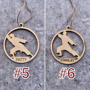 May include: Two wooden Christmas decorations shaped like a karate person with the names "PATTY" and "STANLEY" engraved on them. The decorations are hanging from a string and are numbered #5 and #6.