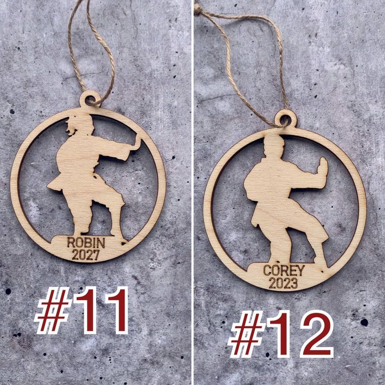 May include: Two wooden Christmas baubles featuring a silhouette of a person in a karate pose. The bauble on the left has the text "Robin 2027" and the number "#11". The bauble on the right has the text "Corey 2023" and the number "#12".