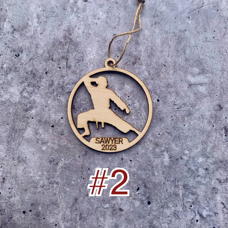 May include: Wooden Christmas bauble featuring a silhouette of a person in a karate pose. The bauble is round and has the text "Sawyer 2023" inscribed on it.
