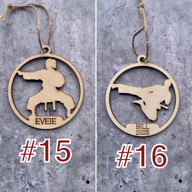 May include: Two wooden Christmas decorations shaped like a person in a karate pose. The decoration on the left has the name "Eveie" written on it and the number "#15" below. The decoration on the right has the name "Bill 2025" written on it and the number "#16" below.