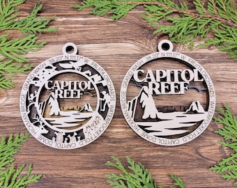 May include: Two wooden ornaments with a layered design featuring the text "CAPITOL REEF". The ornaments include the text "GRAND WASH", "FRUITA ORCHARDS", and "HICKMAN BRIDGE". The ornaments are on a wooden surface with green foliage.