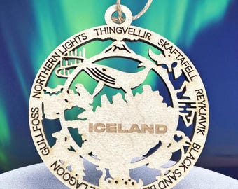May include: A laser-cut wooden ornament featuring the word "ICELAND" and various Icelandic landmarks and phrases, including "Northern Lights," "Thingvellir," and "Blue Lagoon." The ornament has a whale design and is suspended by a natural-colored string.