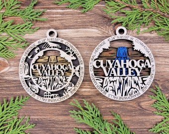 May include: Two wooden ornaments with a laser-cut design of the Cuyahoga Valley National Park. The ornaments are round and feature the text "Cuyahoga Valley" and coordinates. One ornament features a blue waterfall and the other features a brown waterfall.