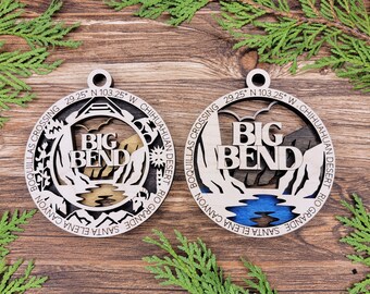 May include: Two wooden ornaments with a laser-cut design of Big Bend National Park in Texas. The ornaments are round and feature the words "Big Bend" in the centre. The top ornament has a gold-coloured river and the bottom ornament has a blue-coloured river.