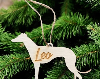 May include: A wooden ornament in the shape of a greyhound dog, with the name "Leo" engraved on its side. The ornament is light-colored and hangs from a natural twine string. The background is a blurred view of green pine needles.