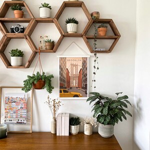 May include: A white wall with a set of hexagonal shelves made of wood. The shelves are decorated with potted plants and a framed print of a city skyline with the word "BROOKLYN" at the top. The shelves are mounted on a white wall above a wooden desk with a vase of dried flowers, a stack of books, and a potted plant.