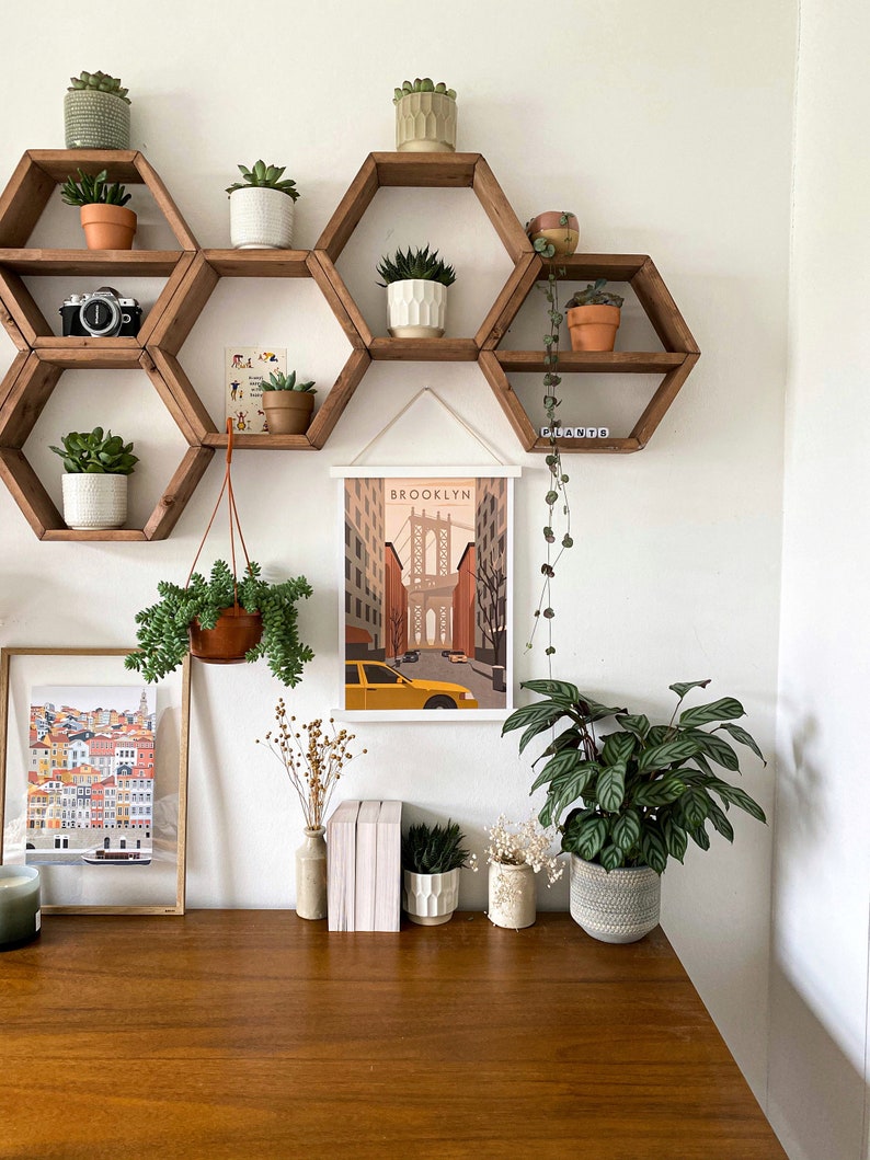 May include: A white wall with a set of hexagonal shelves made of wood. The shelves are decorated with potted plants and a framed print of a city skyline with the word "BROOKLYN" at the top. The shelves are mounted on a white wall above a wooden desk with a vase of dried flowers, a stack of books, and a potted plant.