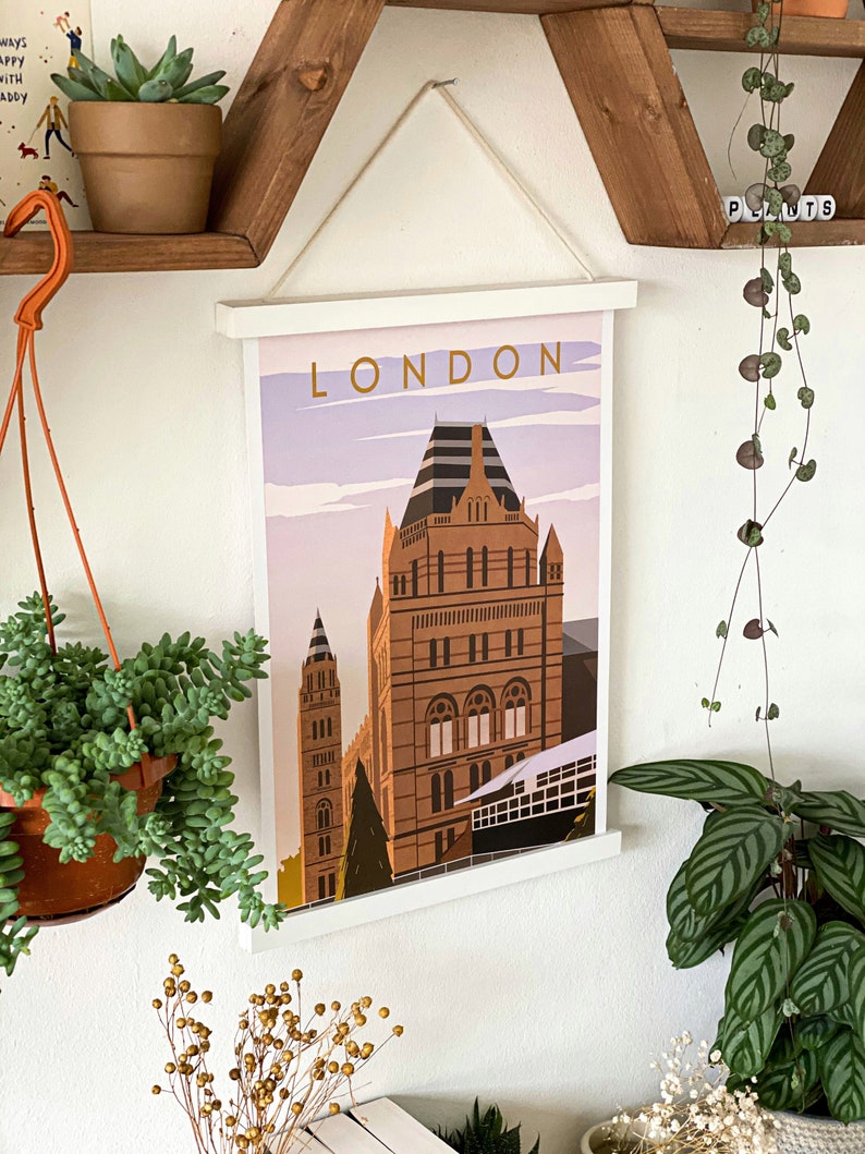 May include: A white framed poster with the word "LONDON" in gold lettering. The poster features a colorful illustration of a building with a pointed roof and a light blue sky.