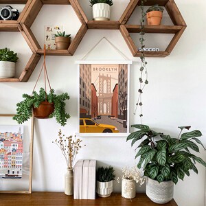 May include: A white framed print with the word "BROOKLYN" and a stylized illustration of the Brooklyn Bridge and a yellow taxi cab. The print is hanging on a wall with a hexagonal wooden shelf unit with plants in pots.