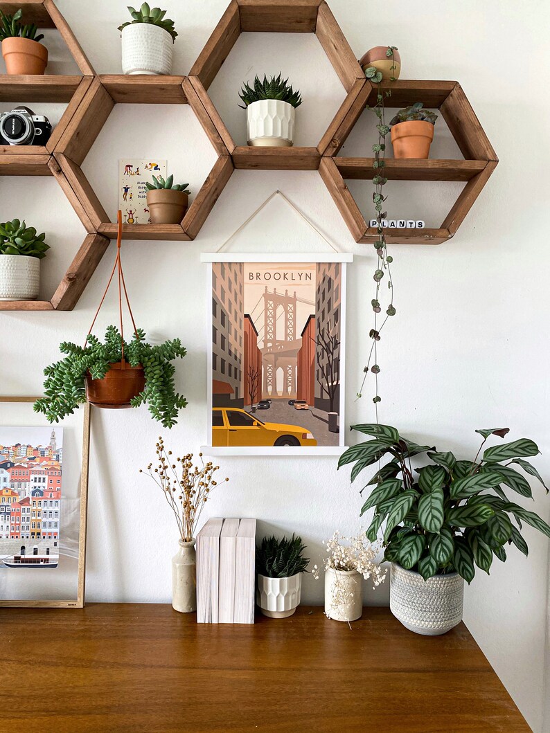 May include: A white framed print with the word "BROOKLYN" and a stylized illustration of the Brooklyn Bridge and a yellow taxi cab. The print is hanging on a wall with a hexagonal wooden shelf unit with plants in pots.