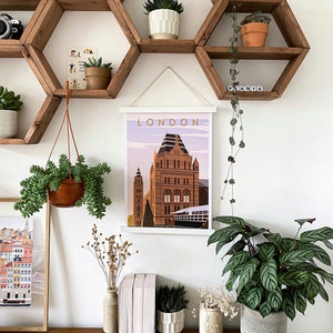 May include: A white framed poster with the word "LONDON" in gold lettering, featuring a colorful illustration of a building with a clock tower. The poster is hanging on a wall with hexagonal wooden shelves, decorated with plants in pots.