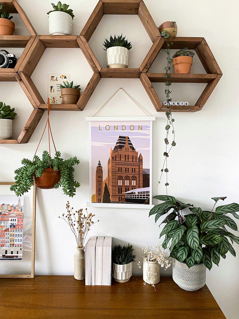 May include: A white framed poster with the word "LONDON" in gold lettering, featuring a colorful illustration of a building with a clock tower. The poster is hanging on a wall with hexagonal wooden shelves, decorated with plants in pots.