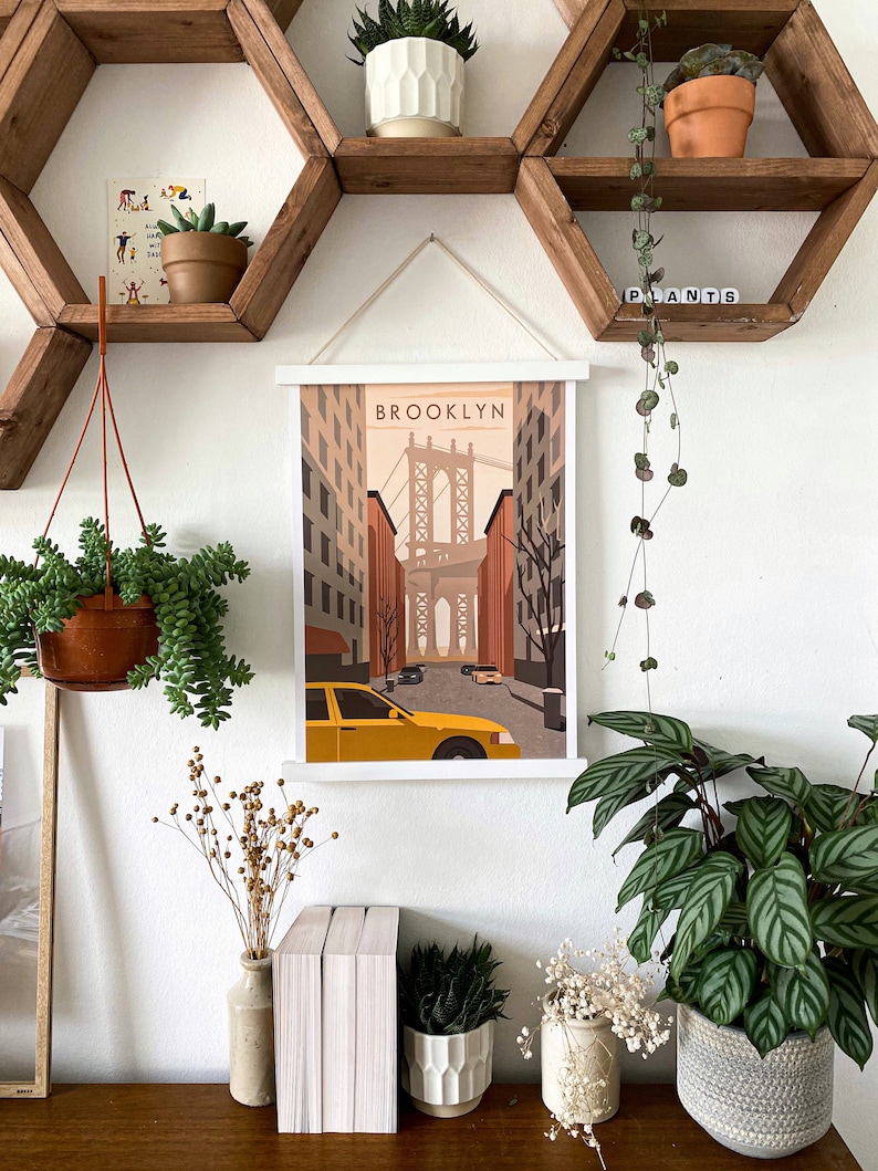 May include: A white framed print with a vintage illustration of the Brooklyn Bridge and the text "BROOKLYN". The print is hanging on a white wall with wooden hexagonal shelves above it. The shelves are decorated with potted plants.