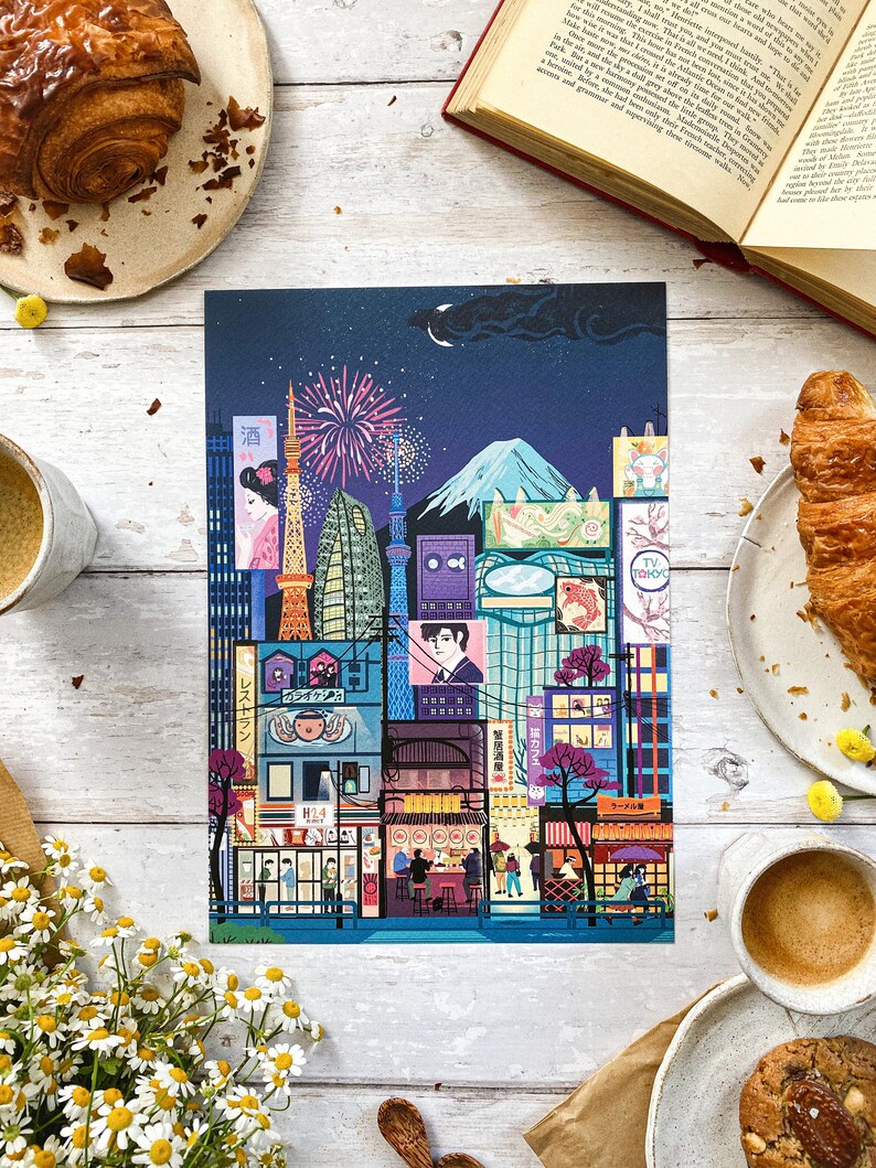 May include: A vibrant print showcasing a stylized Tokyo cityscape at night, with iconic landmarks, fireworks, and traditional buildings. The artwork includes Japanese text and a mountain silhouette. Croissants and coffee cups surround the print.