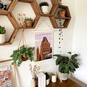 May include: A white framed poster with the word "LONDON" printed in black at the top and a colorful illustration of a building in the center. The poster is hanging on a wall with a hexagonal wooden shelf unit above it. The shelf unit is decorated with potted plants.