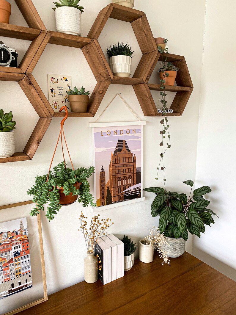 May include: A white framed poster with the word "LONDON" printed in black at the top and a colorful illustration of a building in the center. The poster is hanging on a wall with a hexagonal wooden shelf unit above it. The shelf unit is decorated with potted plants.