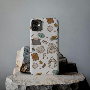Puede incluir: Una funda de teléfono blanca con un patrón repetitivo de ilustraciones que incluye una máquina de escribir, un suéter con motivos de ballenas, un salvavidas con la etiqueta Cabot Cove, libros, gafas, una lupa, una linterna y globos de texto.