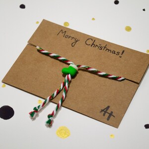May include: A brown paper envelope with a red, white, and green string tied around it. The string has a green heart-shaped charm. The envelope has the words "Merry Christmas!" written on it and the letter "A" followed by a plus sign.