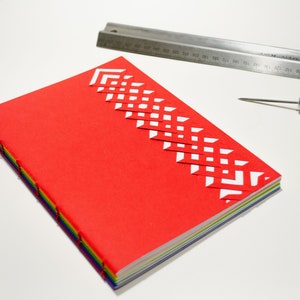 Peut inclure: Un carnet rouge avec un motif géométrique blanc sur la couverture. Le carnet est relié avec du fil de couleur arc-en-ciel et a une colonne vertébrale blanche. Une règle en métal et un poinçon en cuir sont également visibles sur l'image.