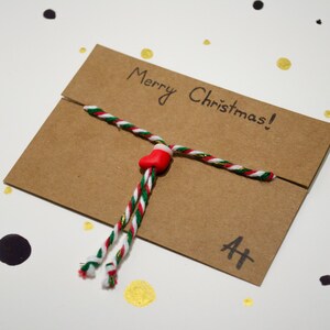 May include: A brown paper envelope tied with a red, white, and green string with a red stocking charm. The envelope has the words "Merry Christmas!" written on it.