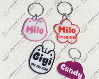 May include: Four colorful personalized pet ID tags with key rings. The tags are shaped like a cat, a flower, a cat, and a cloud. The tags have the names Milo, Gigi, Milo, and Candy, along with phone numbers.