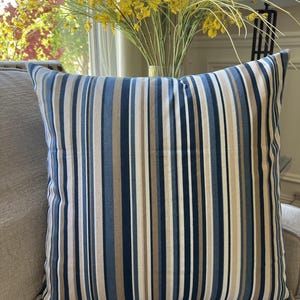 May include: A striped throw pillow with blue, beige, and brown stripes. The pillow is on a light brown couch.