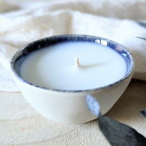 Peut inclure: Une bougie blanche avec une mèche en bois dans un bol en céramique blanche avec un bord bleu.