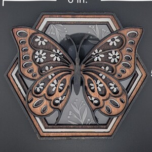 May include: A wooden butterfly wall art piece, 6 inches wide and 5.2 inches tall, with a layered design. The butterfly has brown and black wings with white floral accents, set within a hexagonal frame.