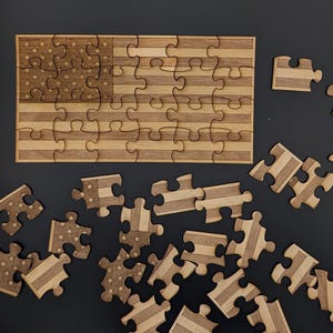 Può includere: Puzzle in legno raffigurante la bandiera americana, con stelle e strisce. Il puzzle è parzialmente assemblato, con pezzi sparsi. Il legno è di colore marrone chiaro naturale e i pezzi sono tagliati con precisione.