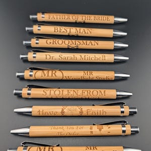 May include: A collection of bamboo pens with silver accents. Each pen is engraved with text such as "Father of the Bride," "Best Man," and "Groomsman." Other pens are engraved with "Dr. Sarah Mitchell," "Stolen From," "Have Faith," and "Thank You For The Order."