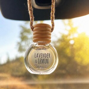 May include: A small, round, clear glass car air freshener bottle with a light wooden cap, suspended by a braided tan cord. The bottle contains a pale yellow liquid and features black text that reads CHILL VIBES LAVENDER & LEMON ESSENTIAL OILS.