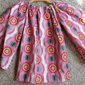 May include: A pink, yellow, and black patterned top with wide sleeves. The fabric features a repeating circular design with a white background. The top is displayed on a wooden hanger.