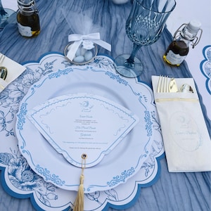 Puede incluir: Una mesa decorada con platos con motivos florales azules y blancos, un menú y una borla dorada. El conjunto incluye una copa de vino azul, pequeñas botellas de aceite de oliva y un paquete de cubiertos. También hay un pequeño recipiente con almendras azucaradas.