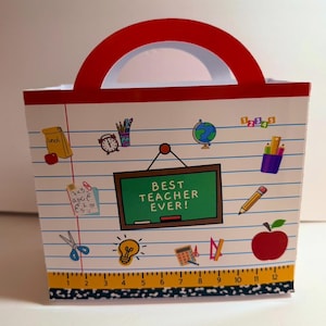 K&ouml;nnte beinhalten: Eine wei&szlig;e Geschenkt&uuml;te mit rotem Griff, verziert mit Aufklebern zum Thema Schule. Die Tasche hat eine gr&uuml;ne Tafel mit der Aufschrift "BEST TEACHER EVER!" und enth&auml;lt Bilder von einem Apfel, einem Bleistift und einem Globus. Der Boden der Tasche hat ein Lineal-Design.