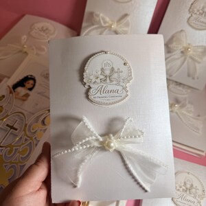 Puede incluir: Tarjetas de invitación blancas con lazos decorativos y detalles de perlas. Las tarjetas presentan un emblema religioso con el nombre "Alana" y el texto "Mi Primera Comunión". Las tarjetas están diseñadas para una celebración de Primera Comunión.