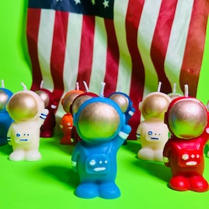 Puede incluir: Una colección de velas con forma de astronauta en varios colores, incluyendo azul, rojo y blanco, con viseras doradas. Las velas están dispuestas frente a una bandera americana sobre un fondo verde. Un artículo decorativo único.