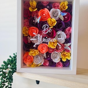 Puede incluir: Una obra de arte floral enmarcada con un borde blanco. La obra presenta flores de papel en tonos naranja, amarillo, morado, rosa y blanco, con detalles de mariposas. Las palabras "Be Unique" se muestran en el centro.