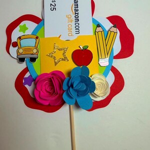 K&ouml;nnte beinhalten: Ein farbenfroher, bl&uuml;tenf&ouml;rmiger Geschenkkartenhalter mit einer 25-$-Amazon-Geschenkkarte. Das Design umfasst einen gelben Schulbus, Bleistifte, einen roten Apfel und Papierblumen in Pink, Blau und Wei&szlig;. Der Geschenkkartenhalter befindet sich auf einem Holzstab.