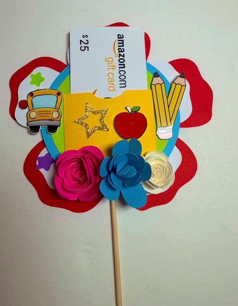 K&ouml;nnte beinhalten: Ein farbenfroher, bl&uuml;tenf&ouml;rmiger Geschenkkartenhalter mit einer 25-$-Amazon-Geschenkkarte. Das Design umfasst einen gelben Schulbus, Bleistifte, einen roten Apfel und Papierblumen in Pink, Blau und Wei&szlig;. Der Geschenkkartenhalter befindet sich auf einem Holzstab.