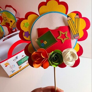 K&ouml;nnte beinhalten: Bunte Papierblumen-Dekoration zum Thema Lehrer. Die Blume hat rote, gelbe und wei&szlig;e Bl&uuml;tenbl&auml;tter, mit einem roten Ordner, einem Stern und Bleistiften in der Mitte. Die Blume ist an einem Holzstab befestigt. Eine passende Geschenkt&uuml;te ist im Hintergrund.