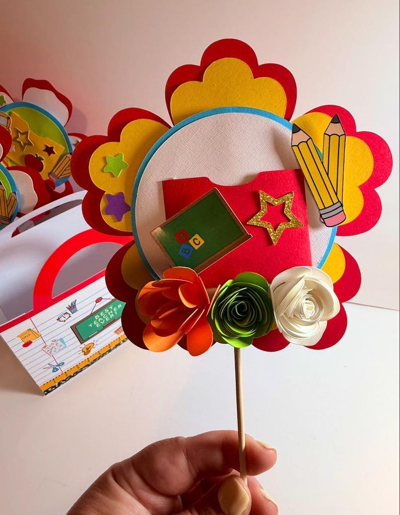 K&ouml;nnte beinhalten: Bunte Papierblumen-Dekoration zum Thema Lehrer. Die Blume hat rote, gelbe und wei&szlig;e Bl&uuml;tenbl&auml;tter, mit einem roten Ordner, einem Stern und Bleistiften in der Mitte. Die Blume ist an einem Holzstab befestigt. Eine passende Geschenkt&uuml;te ist im Hintergrund.