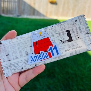Puede incluir: Una barra de chocolate con leche con un envoltorio temático de Snoopy. El envoltorio presenta un diseño de cómic con ilustraciones en blanco y negro. Una pegatina con Snoopy y Woodstock en una caseta roja está en el envoltorio. El texto "Amelia 11" es visible.