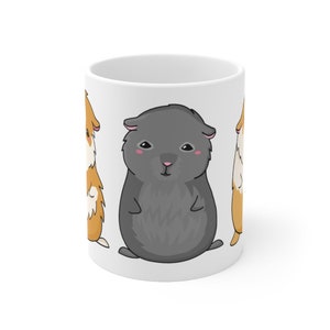 May include: White ceramic mug with a cartoon illustration of three guinea pigs. Two of the guinea pigs are brown and white, and one is gray. The gray guinea pig is in the center of the mug.